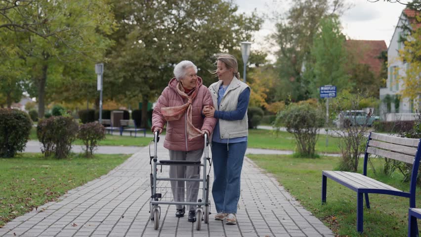 Senior woman and home caregiver spending a chilly, windy day outdoors in city park. Autumn walk for elderly patient with walker.