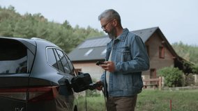 Man charging electric car in front of his house, plugging the charger into the charging port. House with solar panel system on roof behind him. - Powered by Shutterstock - Get 15% off with code: PIKWIZARD15