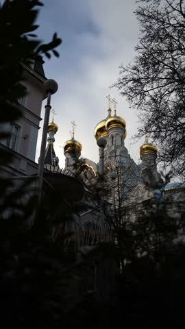 Vertical screen: A serene view of golden domes of an Orthodox church partially hidden by trees, creating a mystical atmosphere. The intricate details of the architecture are framed beautifully by natu