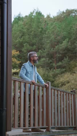 Man is enjoying a relaxing, peaceful moment at home, standing on patio, leaning against railing and looking at autumn nature around him.