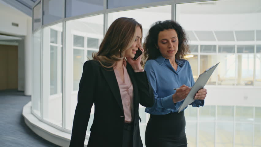 Business colleagues discussing document on go. Smiling woman assisting executive consulting on project. Confident businesswoman talking mobile phone walking office hallway. Corporate people closeup - Powered by Shutterstock - Get 15% off with code: PIKWIZARD15
