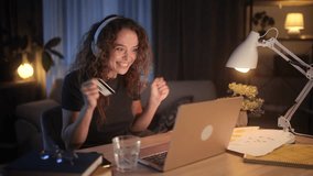 A young woman wearing headphones enters her credit card details and celebrates an online purchase. She smiles excitedly, enjoying shopping from home in a cozy evening setting - Powered by Shutterstock - Get 15% off with code: PIKWIZARD15