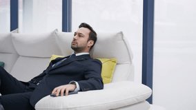 A man in a formal suit reclines relaxing casually on a white sofa with colorful cushions in the background, set against large windows. Businessman relaxing on couch at home smiling - Powered by Shutterstock - Get 15% off with code: PIKWIZARD15