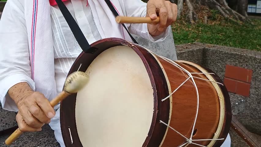 An old Colombian man with a hat and a white shirt playing a drum while sitting on a park bench in Yaguara - Huila - Colombia. Colombian culture and music concept
