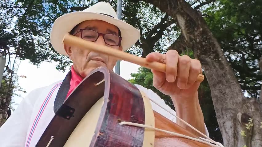 An old Colombian man with a hat and a white shirt playing a drum while sitting on a park bench in Yaguará - Huila - Colombia. Colombian culture and music concept