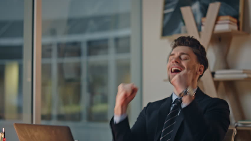 Excited businessman looking laptop reacting emotionally on success at office closeup. Overjoyed man raising hands rejoicing business win at evening workplace. Corporate employee laughing feeling joy.