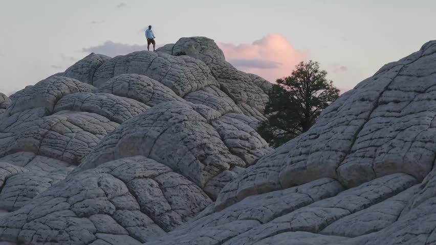 A zoomed-in drone raising altitude to reveal the famous lone tree and a male subject standing on top of the unique sandstone rock features of White Pocket Arizona at sunset with parallax