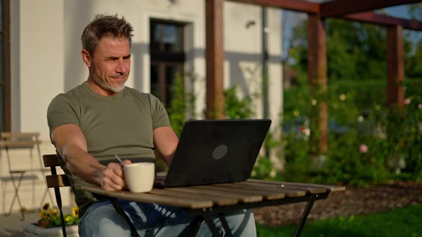 Portrait of happy mature man using laptop drinking coffee outdoor. Businessman working remotely in home office if garden. Smiling male entrepreneur freelancer managing digital business online.