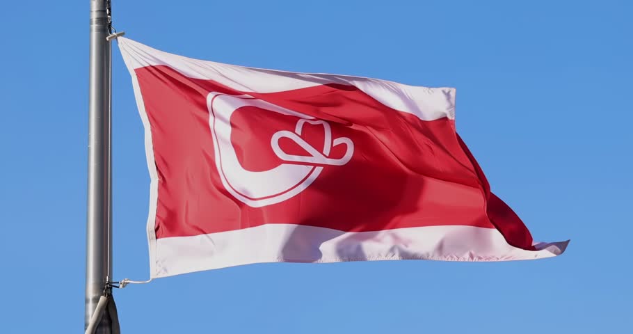 A red and white flag of Calgary with the city's stylized "C" logo featuring a cowboy hat, waving against a clear blue sky.