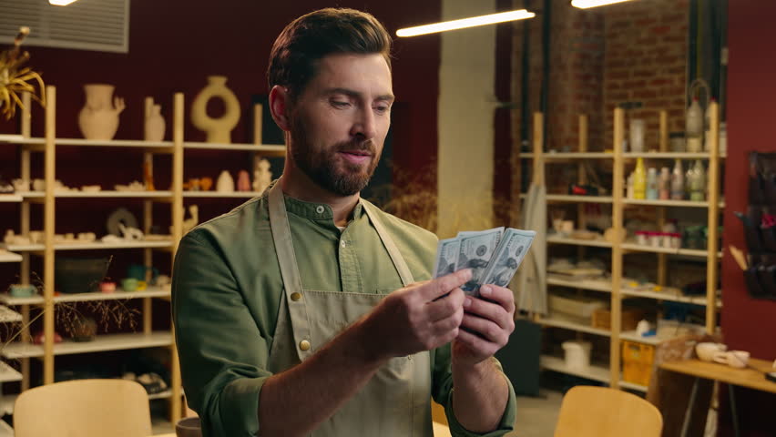 Rich wealthy happy Caucasian man male businessman guy small business owner entrepreneur potter apron counting money dollars winning cash salary finance wage budget payment wealth inside pottery store