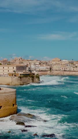 Vertical aerial drone shot of Ortigia, Syracuse. Flying along the coast of Ortigia island with large waves crashing on the shore. Historic Italian town in Sicily.