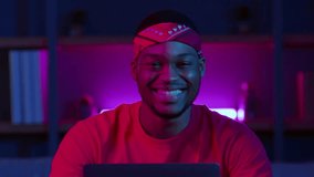 A man smiles while working on his laptop in a stylish home environment. Neon lights create a vibrant atmosphere, emphasizing the modern, tech-savvy lifestyle of a digital nomad at night. - Powered by Shutterstock - Get 15% off with code: PIKWIZARD15
