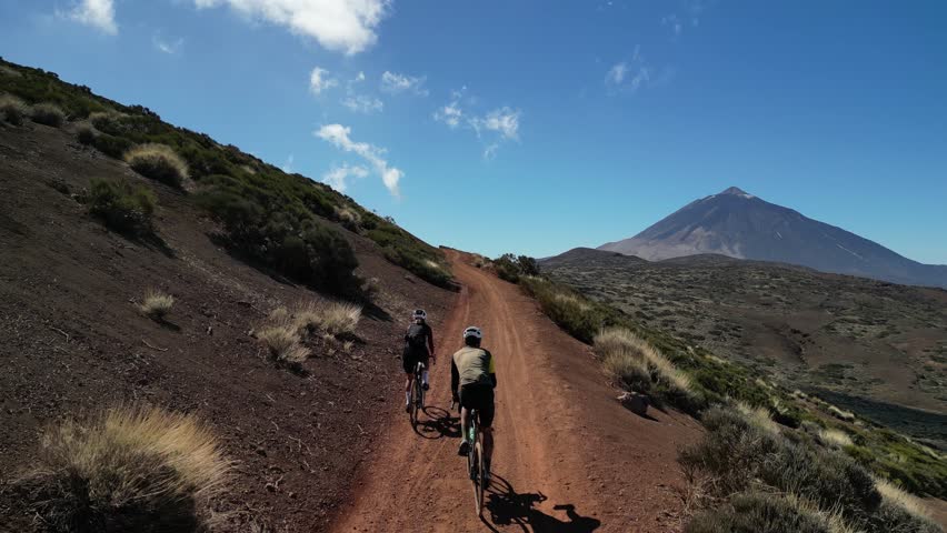 Two cyclists ride along a scenic red dirt road. The vast landscape under a bright blue sky provides a stunning backdrop for this active outdoor adventure.Teide. Tenerife. Spain
