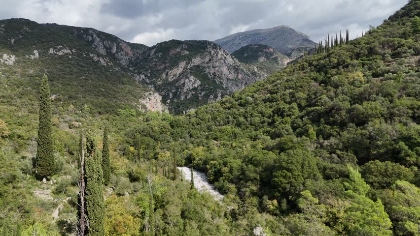 Aerial drone flight through Vyros Gorge near Kardamyli, Greece, on a sunny winter day. The gorge features lush green vegetation, striking red rock formations, and surrounding mountains.