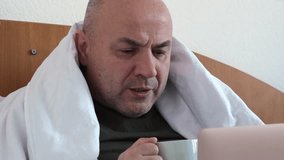 Sick middle aged man sitting on bed in front of laptop with cup of hot tea and coughing. Employee is on a video call, close up. Person is working from home despite illness - Powered by Shutterstock - Get 15% off with code: PIKWIZARD15