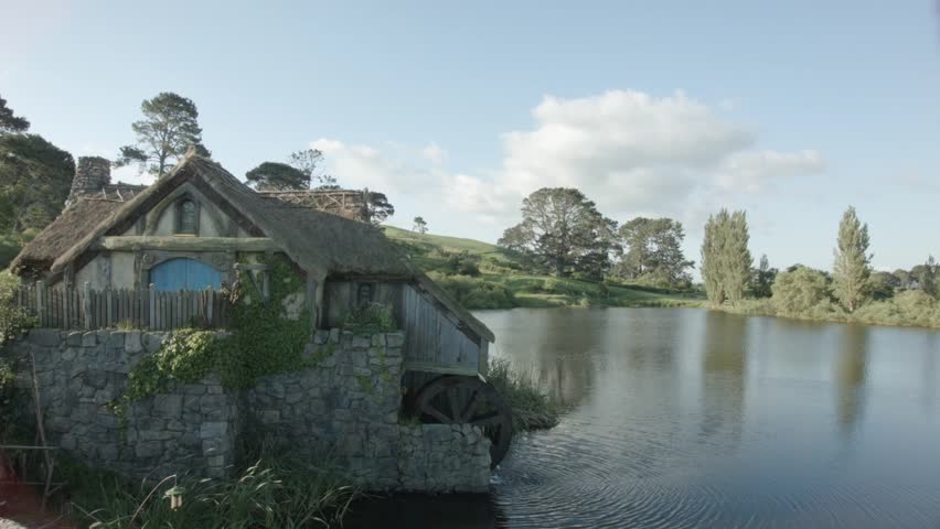 A serene countryside scene with a rustic watermill by a tranquil lake, surrounded by lush greenery and rolling hills under a clear blue sky.