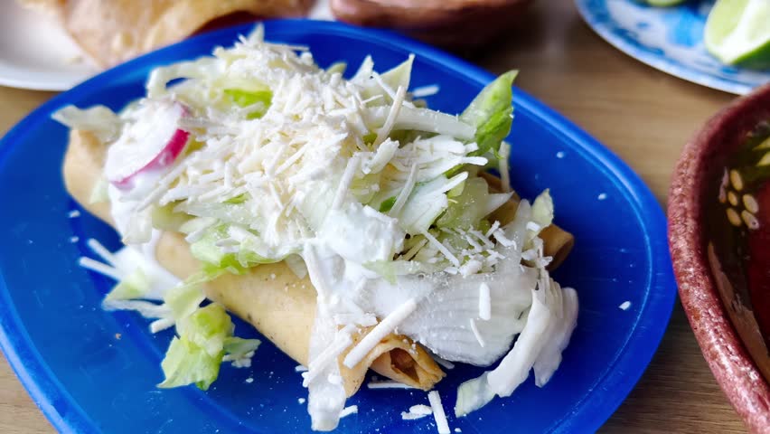 Delicious flautas, typical mexican food, lying on a blue plate and covered with shredded cheese, cream, lettuce and radish slices, ready to be eaten