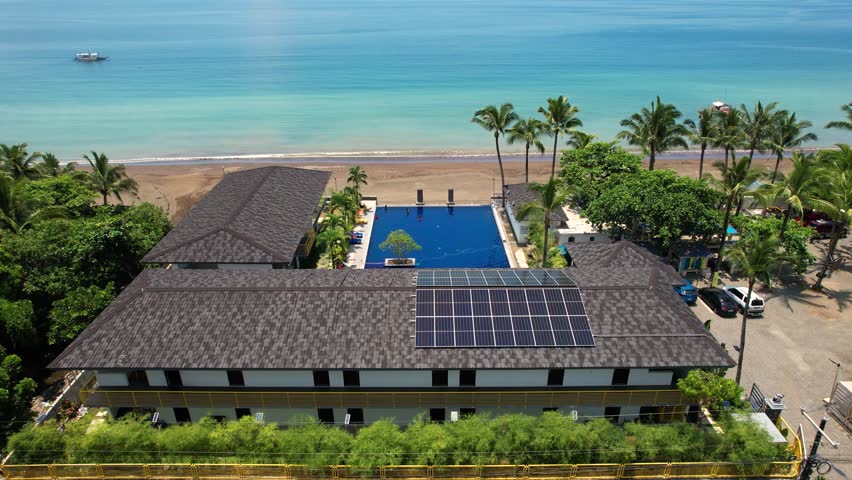 An array of Solar panels mounted on a resort building rooftop, with a beautiful tropical beach and palm trees in the background. At Real, Quezon, Philippines.