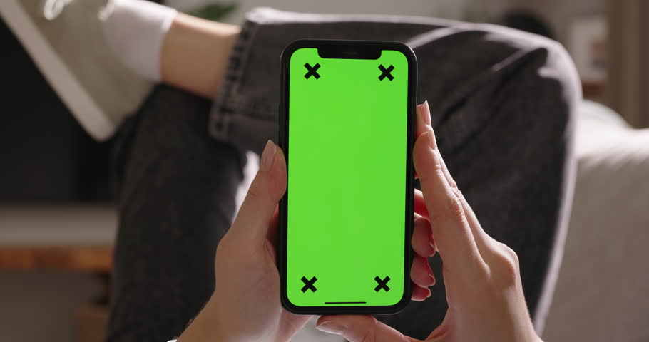 Young woman holds phone with green screen while lounging on sofa, vertical orientation. Advertising app close-up. Tap in center to immersed in captivating video content. World of endless possibilities