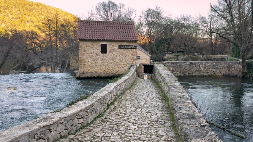 Beautiful woman in grey coat walking over stone bridge toward an old stone watermill in Krka National Park, Croatia