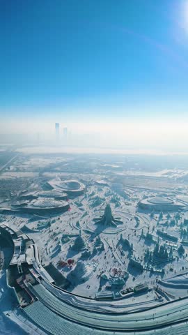 Vertical aerial view of the Harbin Ice Festival 2025, showcasing intricate ice sculptures, frozen palaces, and a vast winter wonderland set against the city skyline under clear blue skies. China