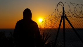 Hooded figure contemplating sunset behind barbed wire. Solitary hooded silhouette standing beside barbed wire fence, gazing across golden sunset, themes of freedom, solitude, and unspoken journey - Powered by Shutterstock - Get 15% off with code: PIKWIZARD15