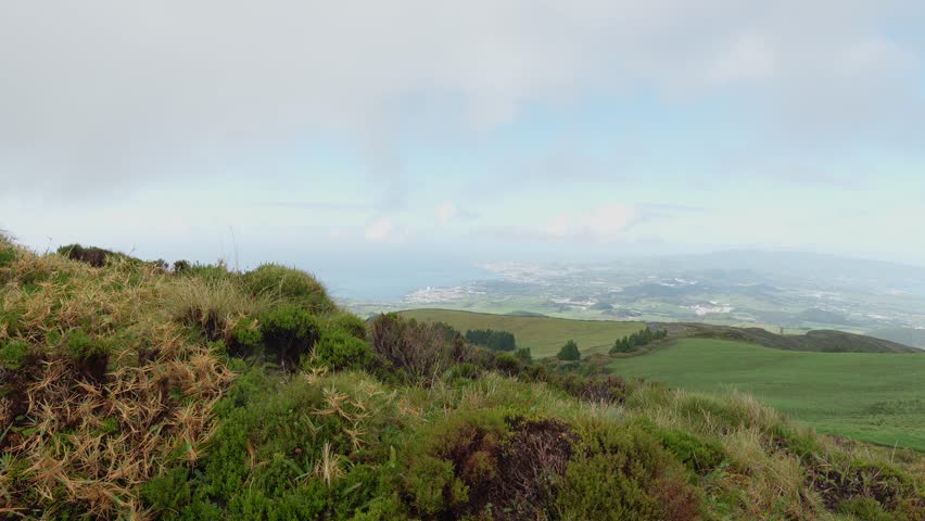 A view of green grassy hillside under cloudy sky with vast fields in the background at the Sao Miguel island, the Azores, Portugal