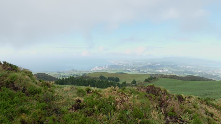 A view of green grassy hillside under cloudy sky with vast fields in the background at the Sao Miguel island, the Azores, Portugal