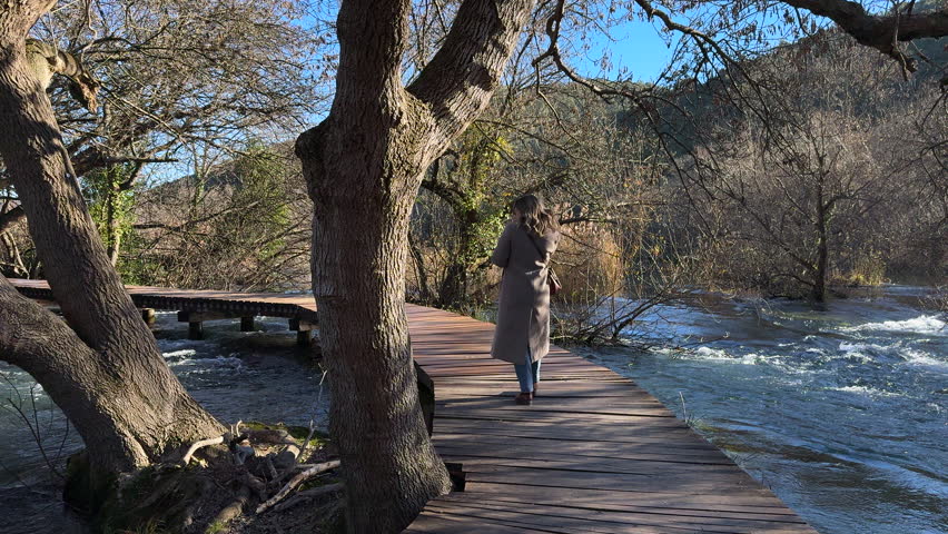 Beautiful woman in grey coat walking on a wooden bridge over a water stream in Krka National Park, Croatia