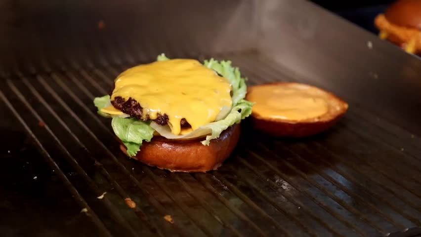 Cooking process A chef at the grill adds crispy bacon to a burger with cheese and lettuce. Perfect for video content about cooking, food blogs or restaurant menus.