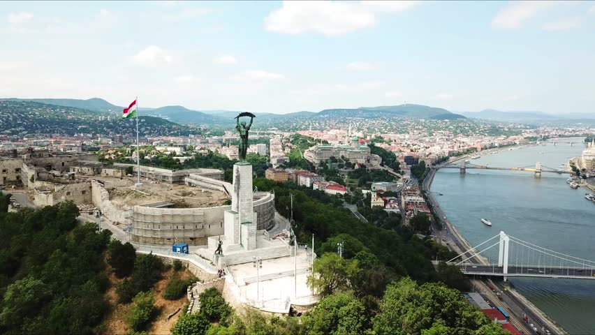 Aerial view of the liberty statue and the citadel on Gellért hill in Budapest, Hungary, showcasing the cityscape and the danube river on a sunny day with the hungarian flag waving, drone orbiting shot