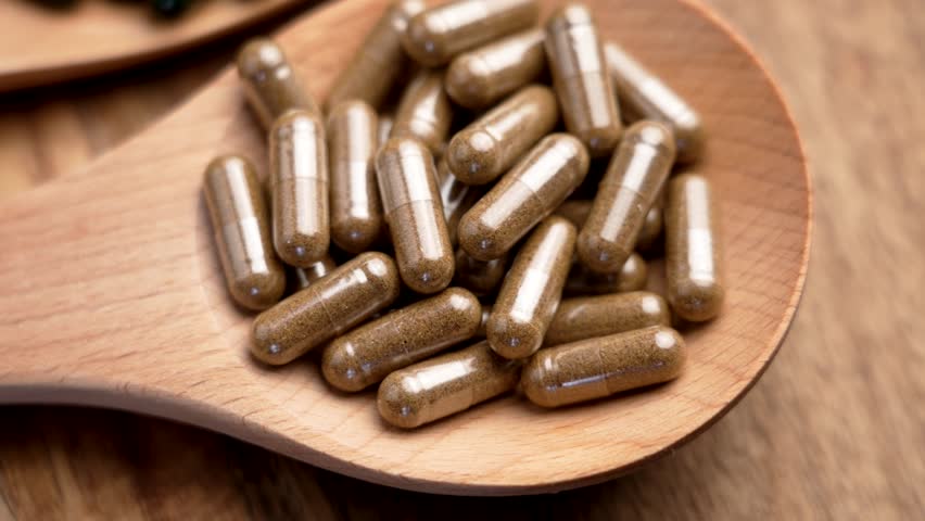 Brown herbal capsules in wooden spoon. Natural plant-based remedies. Close-up shot. Top view. Rotation