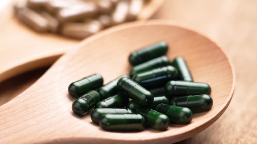 Green herbal capsules falling into a wooden spoon. Assortment of alternative medicine concept. Slow motion