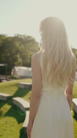 Elegant woman with long blonde hair in a white dress standing outdoors in sunlight, evoking feelings of grace and sophistication. The natural light highlights her serene presence. Woman in White Dress