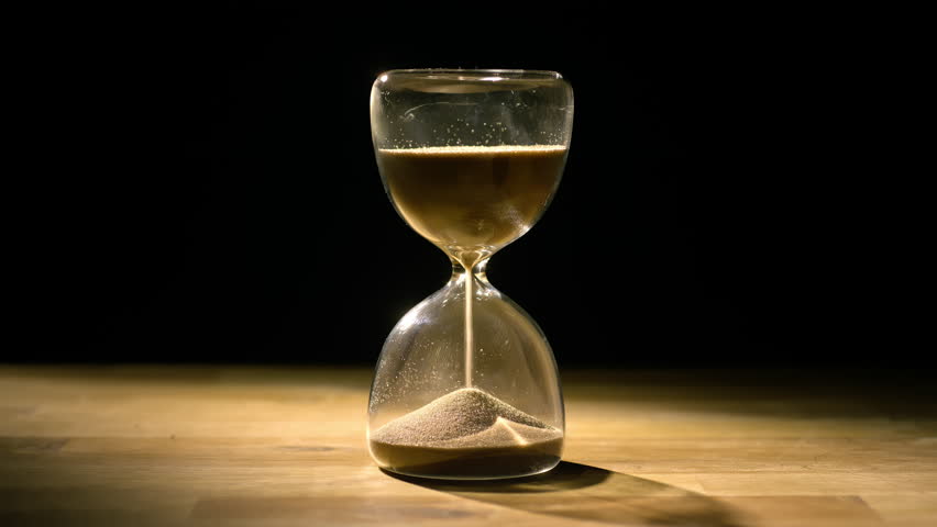 Hourglass on a wooden table and black background, sand passing from the upper to the lower bulb, close up shot. Sands of time concept.