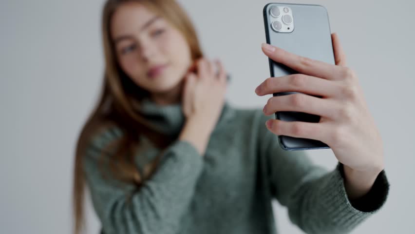 Blurred focus image of young woman in green sweater capturing a selfie with her smartphone in a bright indoor setting. Young Woman in Green Sweater Taking Selfie with Smartphone