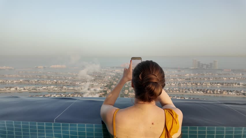 A girl stands on the edge of a pool and looks at a huge city. The pool is on the roof of a skyscraper.