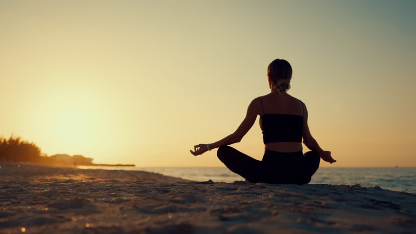 Woman practicing meditation in sandy ocean beach in sunrise time, back view. Relaxation and recovery vitality, healthy lifestyle for stay beautiful, trip to sea in summer vacation, old yoga practice