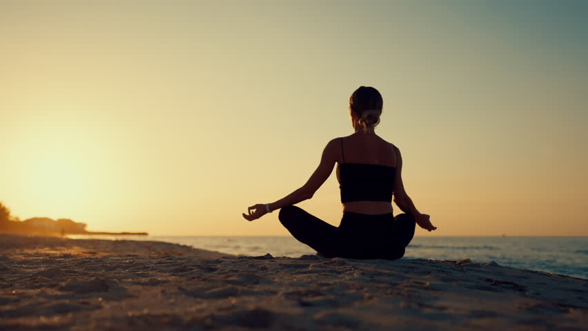 Woman practicing meditation in sandy ocean beach in sunrise time, back view. Relaxation and recovery vitality, healthy lifestyle for stay beautiful, trip to sea in summer vacation, old yoga practice