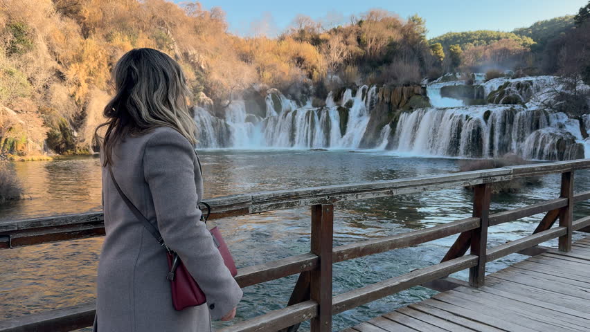 Woman in grey coat walking on a wooden bridge and watching the beautiful waterfalls in Krka National Park, Croatia