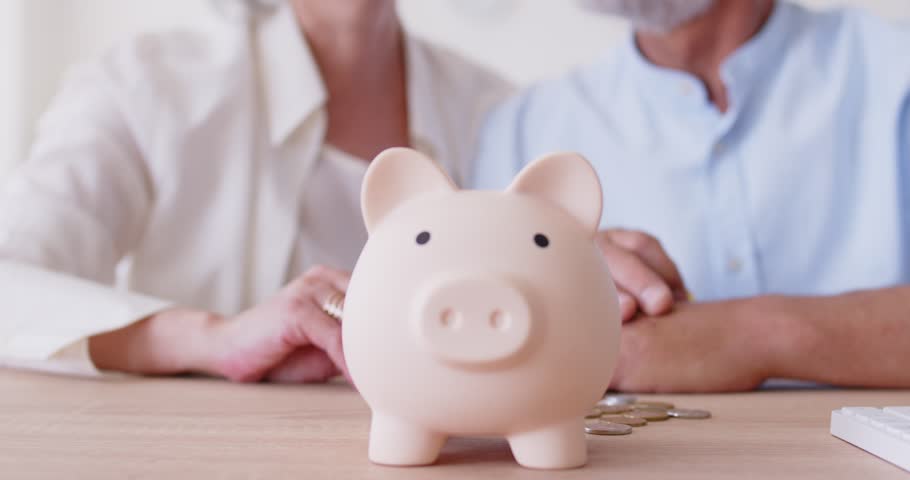 An elderly couple puts coins into a pink piggy bank, hands close-up. Older people save money. Retirement savings, investments for old age. Concept collecting coins, investment banking