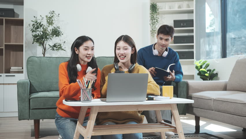 Three Asian young adult students sit in university living room, discussing their study abroad experiences, academic research, preparing thesis report, planning future global education opportunities.