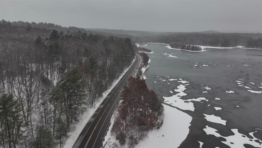 Frozen Lake in suburb of american town. White car driving on road during snowstorm. Fir trees and leafless trees in background. Aerial wide shot.