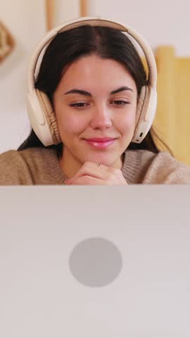 Vertical HD video, front view of happy young student wearing headphones and using laptop for online learning, enjoying breakfast in kitchen. Education and e-learning concept