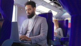 An African American businessman is focused on his laptop while traveling on the train, while a woman in the background uses her phone, highlighting modern multitasking and urban commuting lifestyle - Powered by Shutterstock - Get 15% off with code: PIKWIZARD15