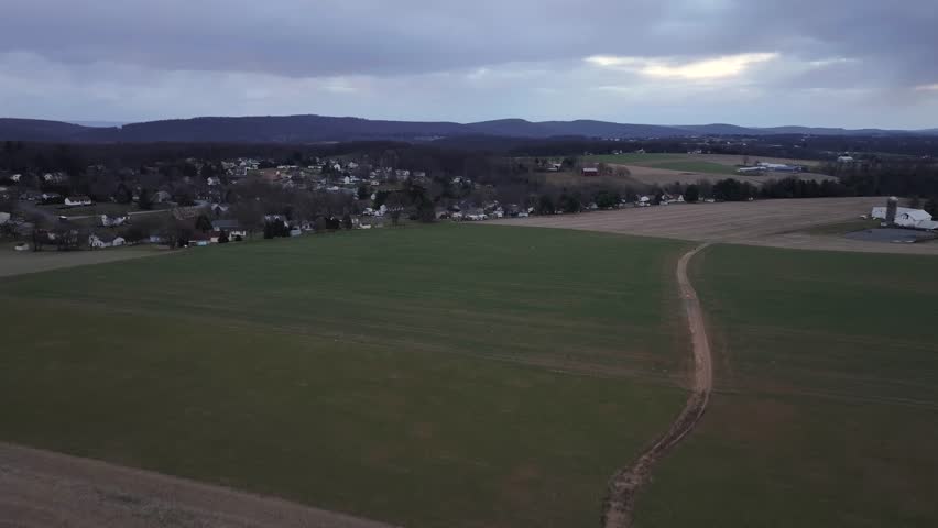 Suburb neighborhood of american town during cloudy winter day. Rising drone wide shot. Agricultural farm fields in January. Silhouette of mountain range in distance.