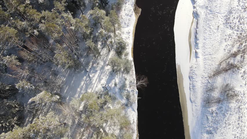 Aerial view of a winding river through a dense snow-covered forest in winter, with a curving road running alongside. A serene and scenic natural landscape.