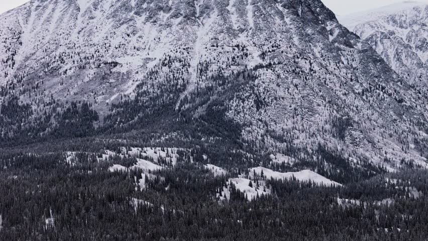 Cinematic aerial view of the imposing snow-covered mountains and dense forests surrounding the Takhini River and Kusawa Lake in Yukon, Canada. A breathtaking winter wilderness scene.