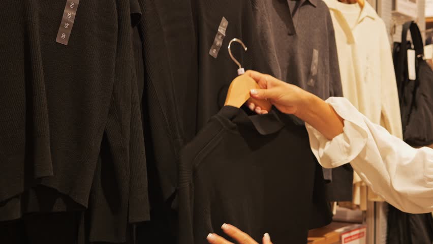 Woman browsing clothing rack in fashion store carefully selecting sweater Stylish garments on wooden hangers warm lighting creates cozy shopping atmosphere making decision on fashion purchase 