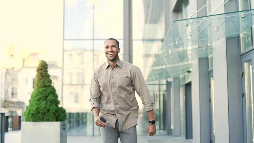 Excited handsome employee jumps and rejoices get good news great result looking at mobile phone. Young adult business male celebrating success walking near office building outside browsing smartphone